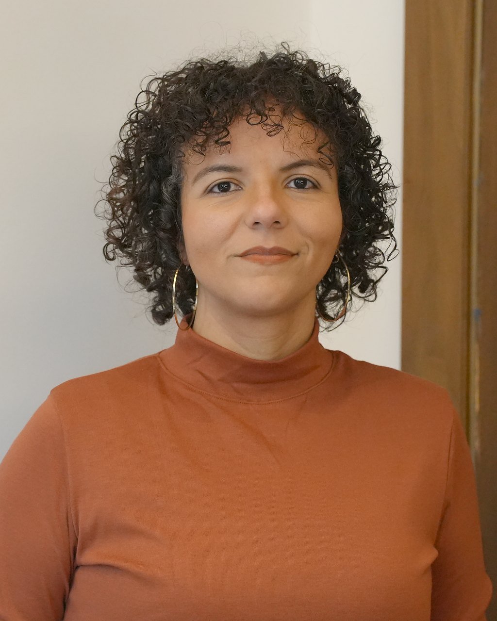 Raquel Camargo looking at the camera, wearing a terracotta top against a plain wall
