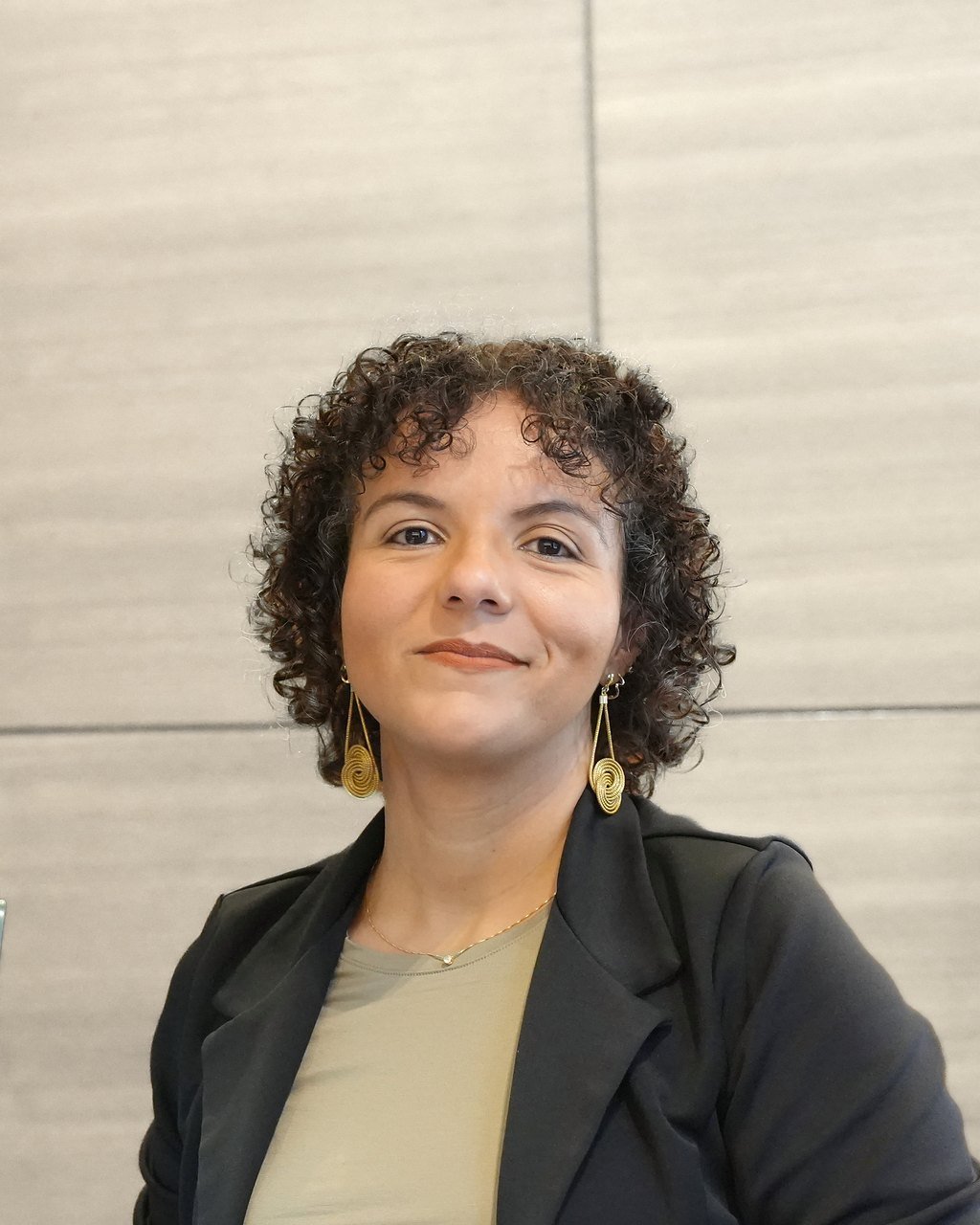 Raquel Camargo holding a laptop, looking at the camera, against a pale straw-coloured wall