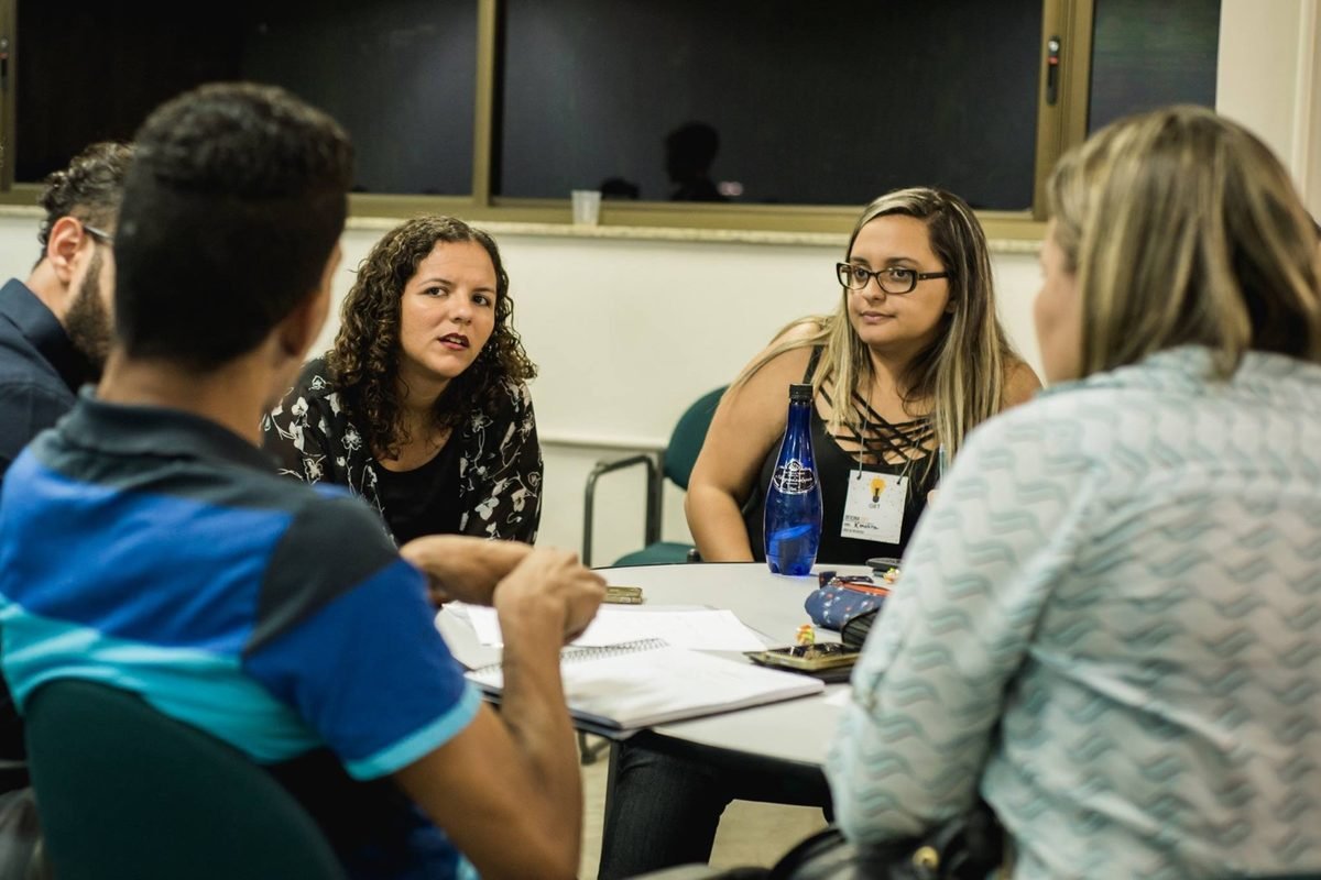 Raquel Camargo talking with three participants around a classroom table, notebooks open between them
