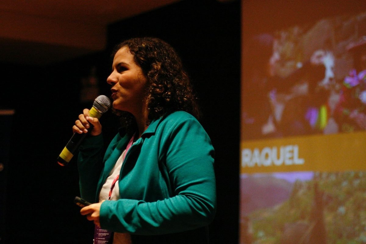 Raquel Camargo on a conference stage with a microphone, her first name projected on the screen behind her