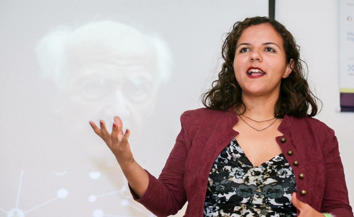 Raquel Camargo mid-sentence in front of a projected slide, one hand raised as she explains a point