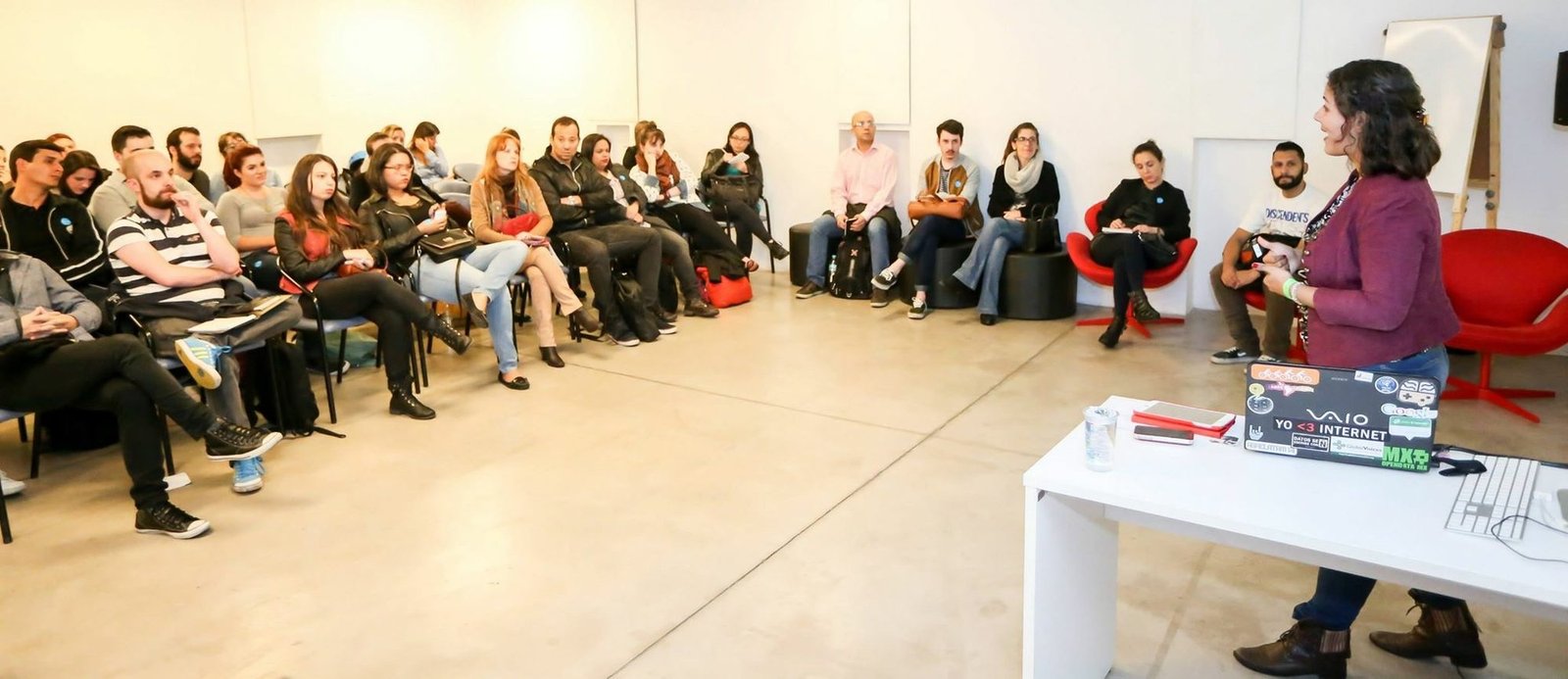 Raquel Camargo standing at the front of a bright studio space, speaking to around twenty-five people seated in rows and on low stools