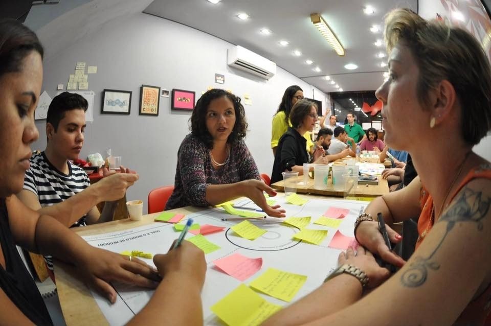 Raquel Camargo at a workshop table covered in post-it notes and a printed canvas, talking with the participants around it